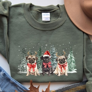 May include: A green Christmas sweatshirt with a design of three pugs wearing Santa hats and reindeer antlers. The pugs are standing in front of a snowy forest scene.