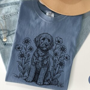 Pode incluir: Uma camiseta azul jeans com uma ilustra&ccedil;&atilde;o em preto e branco de um cachorro sentado entre flores. A camisa est&aacute; dobrada sobre um par de jeans azul claro, com um colar dourado e um chap&eacute;u bege.