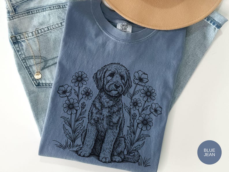 Pode incluir: Uma camiseta azul jeans com uma ilustra&ccedil;&atilde;o em preto e branco de um cachorro sentado entre flores. A camisa est&aacute; dobrada sobre um par de jeans azul claro, com um colar dourado e um chap&eacute;u bege.