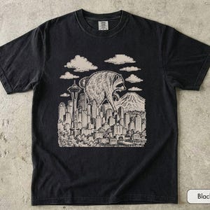 Pu&ograve; includere: Una t-shirt nera a maniche corte con una stampa grafica color crema raffigurante un procione gigante che sovrasta lo skyline di una citt&agrave;. L'illustrazione include lo Space Needle, numerosi grattacieli, nuvole e una montagna sullo sfondo.