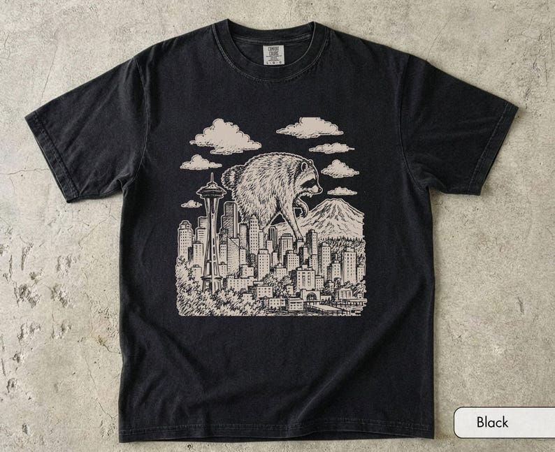 Pu&ograve; includere: Una t-shirt nera a maniche corte con una stampa grafica color crema raffigurante un procione gigante che sovrasta lo skyline di una citt&agrave;. L'illustrazione include lo Space Needle, numerosi grattacieli, nuvole e una montagna sullo sfondo.