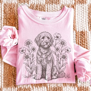 Pode incluir: Uma camisa rosa claro de manga comprida com gola redonda, apresentando uma ilustra&ccedil;&atilde;o em preto e branco de um c&atilde;o sentado entre flores. A palavra "Blossom" est&aacute; em um c&iacute;rculo rosa no canto inferior direito.