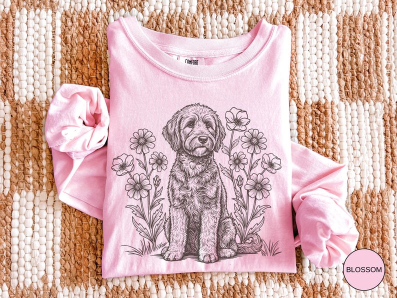 Pode incluir: Uma camisa rosa claro de manga comprida com gola redonda, apresentando uma ilustra&ccedil;&atilde;o em preto e branco de um c&atilde;o sentado entre flores. A palavra "Blossom" est&aacute; em um c&iacute;rculo rosa no canto inferior direito.