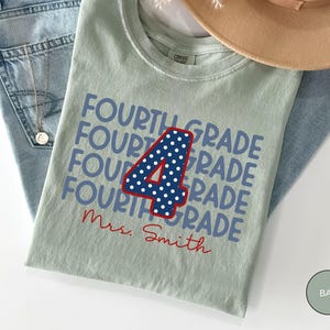 May include: A sage green t-shirt featuring the repeated text "FOURTH GRADE" in blue, a large central number "4" in navy blue with white polka dots and a red outline, and the red cursive text "Mrs. Smith" printed below the design.