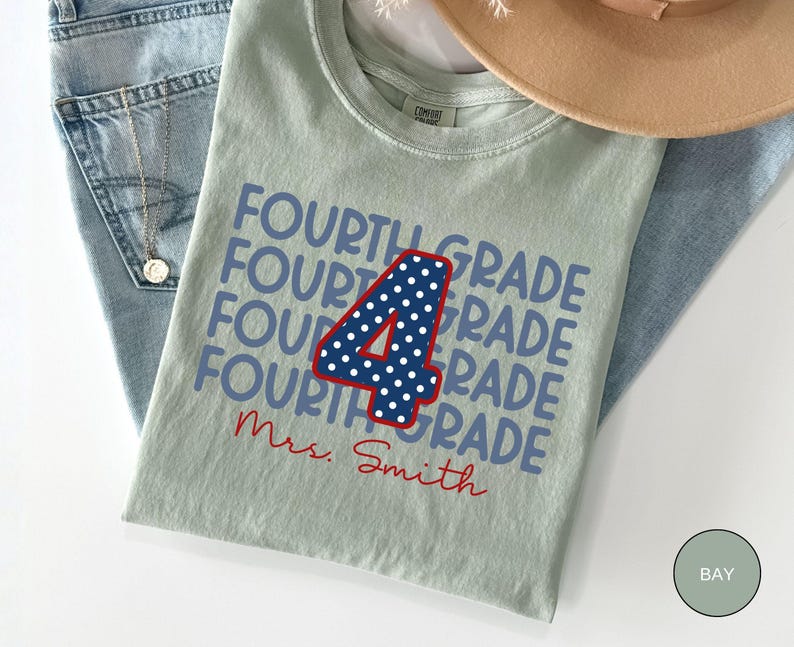 May include: A sage green t-shirt featuring the repeated text "FOURTH GRADE" in blue, a large central number "4" in navy blue with white polka dots and a red outline, and the red cursive text "Mrs. Smith" printed below the design.