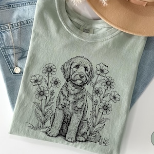 Pode incluir: Camiseta verde claro com uma ilustra&ccedil;&atilde;o em preto e branco de um cachorro sentado entre flores. A camisa est&aacute; dobrada sobre um par de jeans azuis. Um colar dourado e um chap&eacute;u bege tamb&eacute;m s&atilde;o vis&iacute;veis.