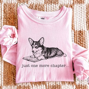 Op de afbeelding: Een lichtroze shirt met lange mouwen met een afbeelding van een corgi-hond die een boek leest. De tekst "just one more chapter..." staat eronder. Het shirt is opgevouwen op een geweven kleed.