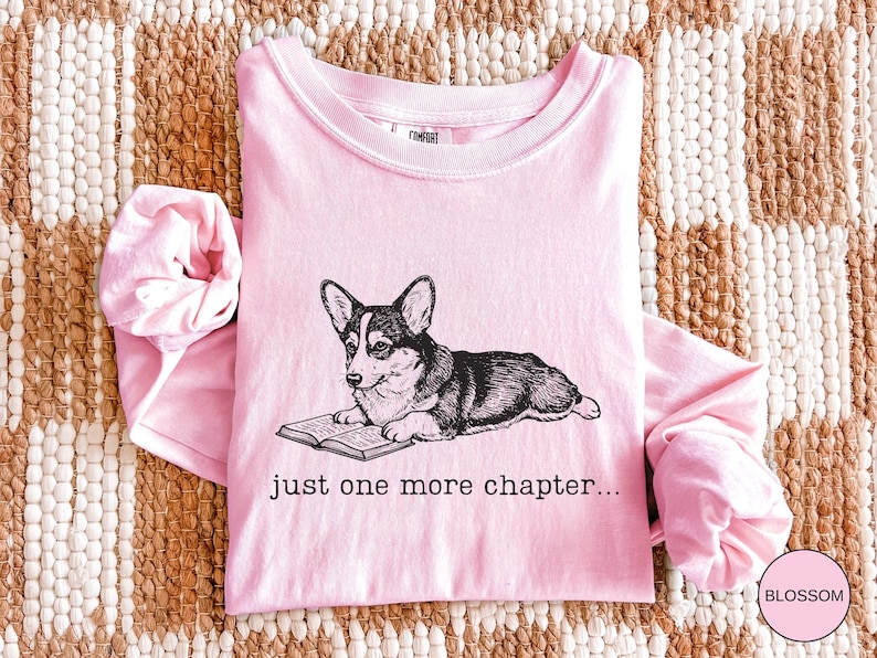 Op de afbeelding: Een lichtroze shirt met lange mouwen met een afbeelding van een corgi-hond die een boek leest. De tekst "just one more chapter..." staat eronder. Het shirt is opgevouwen op een geweven kleed.