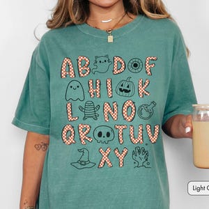 May include: A light green t-shirt featuring a graphic alphabet design where letters are filled with an orange and white checkered pattern. The design includes black line art illustrations of ghosts, a pumpkin, a skull, a witch hat, and a hand.
