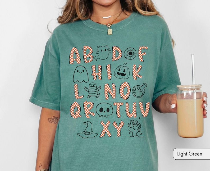 May include: A light green t-shirt featuring a graphic alphabet design where letters are filled with an orange and white checkered pattern. The design includes black line art illustrations of ghosts, a pumpkin, a skull, a witch hat, and a hand.