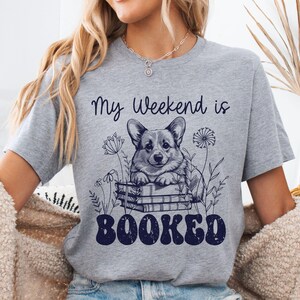 Puede incluir: Camiseta gris con un gr&aacute;fico de un perro corgi sentado en una pila de libros. El texto "My weekend is booked" est&aacute; impreso en la camiseta.