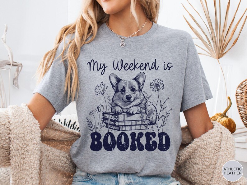 Puede incluir: Camiseta gris con un gr&aacute;fico de un perro corgi sentado en una pila de libros. El texto "My weekend is booked" est&aacute; impreso en la camiseta.
