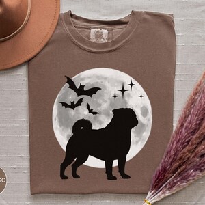 K&ouml;nnte beinhalten: Espressofarbenes T-Shirt mit einer schwarzen Silhouette eines Mops vor einem Vollmond, Flederm&auml;usen und Sternen. Das Shirt hat einen Rundhalsausschnitt und ein Etikett am Kragen. Ein brauner Hut und Trockenblumen sind im Hintergrund.