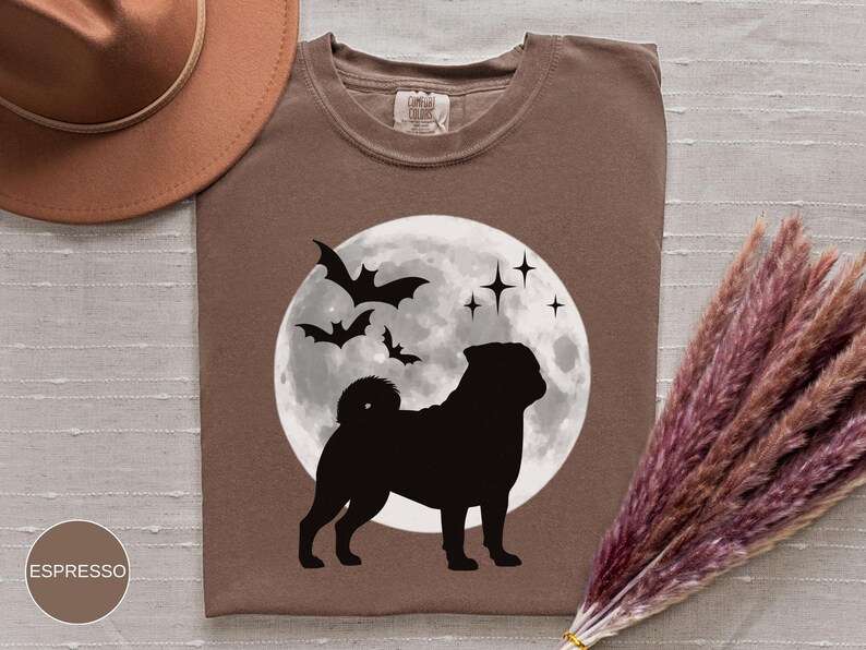 K&ouml;nnte beinhalten: Espressofarbenes T-Shirt mit einer schwarzen Silhouette eines Mops vor einem Vollmond, Flederm&auml;usen und Sternen. Das Shirt hat einen Rundhalsausschnitt und ein Etikett am Kragen. Ein brauner Hut und Trockenblumen sind im Hintergrund.
