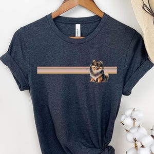 Puede incluir: Camiseta gris oscuro jaspeada con una ilustraci&oacute;n de un perro pomerania marr&oacute;n y negro. El perro est&aacute; sentado frente a un dise&ntilde;o a rayas con tres rayas horizontales en tonos marrones y rosados.