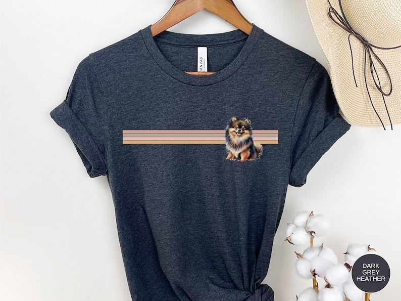 Puede incluir: Camiseta gris oscuro jaspeada con una ilustraci&oacute;n de un perro pomerania marr&oacute;n y negro. El perro est&aacute; sentado frente a un dise&ntilde;o a rayas con tres rayas horizontales en tonos marrones y rosados.