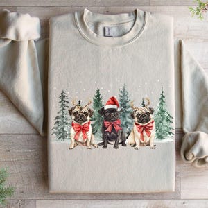 May include: A beige sweatshirt with a Christmas-themed design featuring three pugs wearing Santa hats and red bows. The pugs are standing in front of a snowy forest scene.