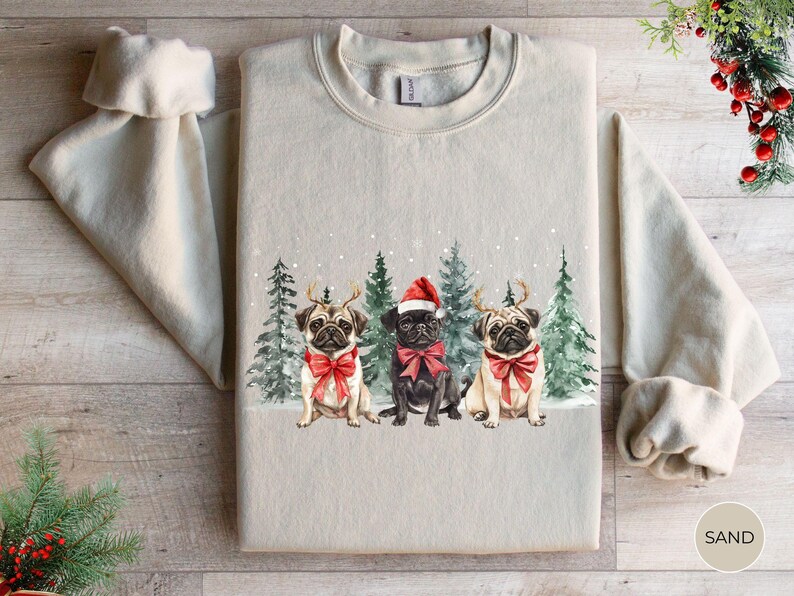 May include: A beige sweatshirt with a Christmas-themed design featuring three pugs wearing Santa hats and red bows. The pugs are standing in front of a snowy forest scene.
