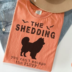 May include: Melon-colored t-shirt with the text "THE SHEDDING" and a silhouette of a dog. The phrase "you can't escape the FLUFF..." is printed below the dog. The shirt is laid on top of blue jeans and a tan hat.
