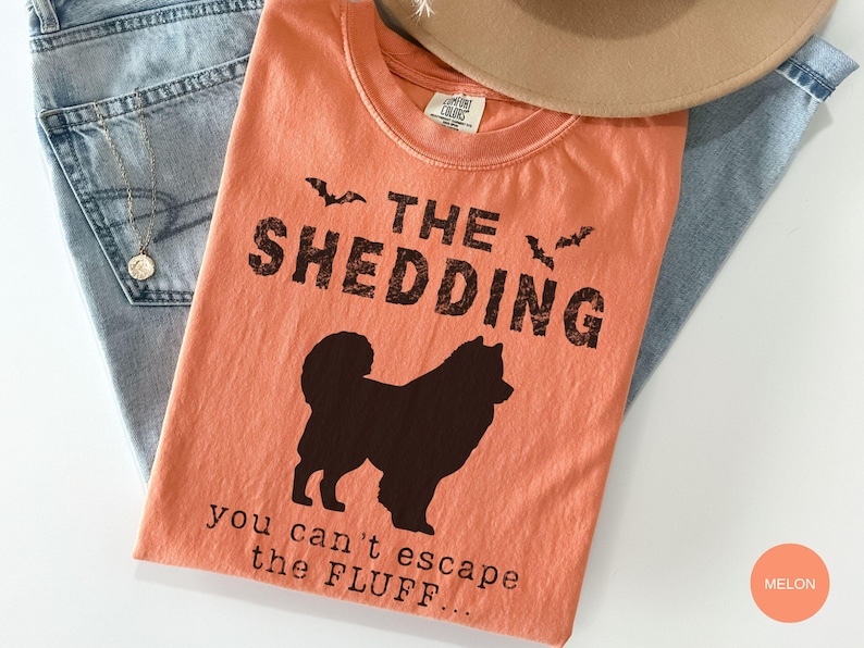 May include: Melon-colored t-shirt with the text "THE SHEDDING" and a silhouette of a dog. The phrase "you can't escape the FLUFF..." is printed below the dog. The shirt is laid on top of blue jeans and a tan hat.