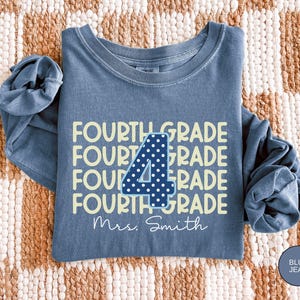 May include: A blue jean colored long-sleeve crewneck sweatshirt featuring the repeating yellow text FOURTH GRADE. A large dark blue number 4 with white polka dots is centered over the text, and the white script text Mrs. Smith is below.