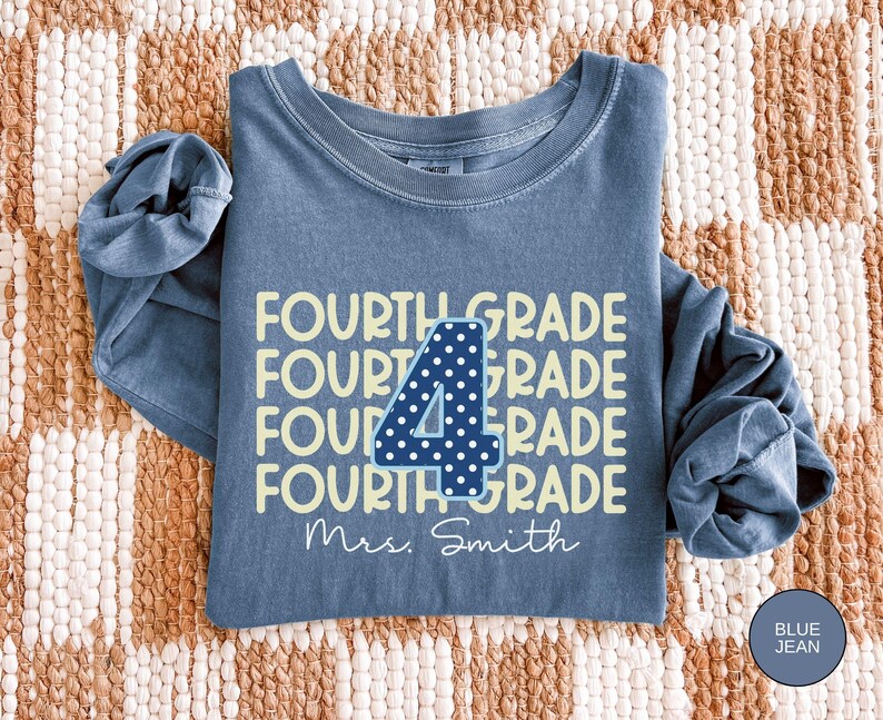 May include: A blue jean colored long-sleeve crewneck sweatshirt featuring the repeating yellow text FOURTH GRADE. A large dark blue number 4 with white polka dots is centered over the text, and the white script text Mrs. Smith is below.
