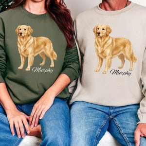 May include: Two sweatshirts, one olive green and one beige, each featuring a golden retriever illustration with the name "Murphy" in script. The dog is depicted in a standing pose. Both individuals are wearing blue jeans.