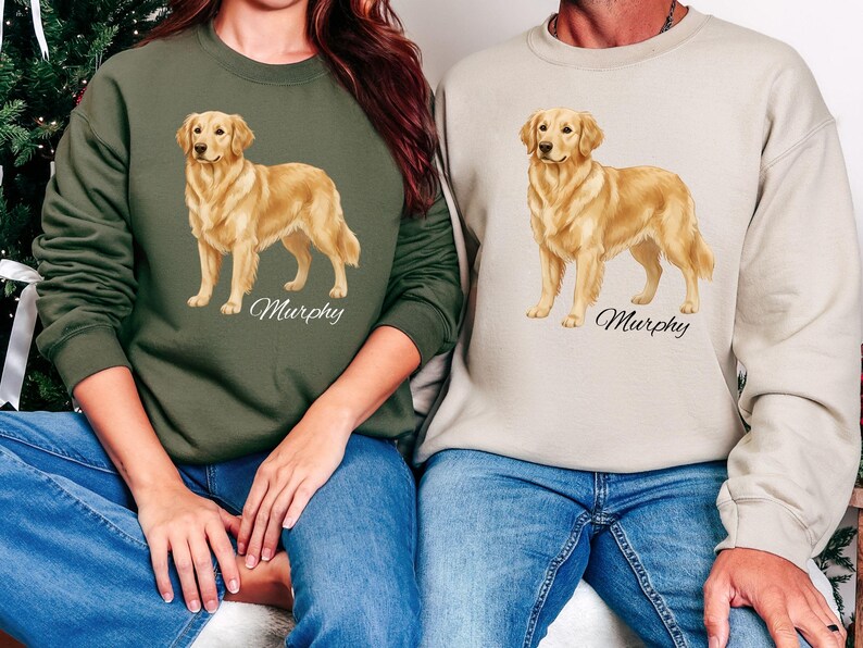 May include: Two sweatshirts, one olive green and one beige, each featuring a golden retriever illustration with the name "Murphy" in script. The dog is depicted in a standing pose. Both individuals are wearing blue jeans.