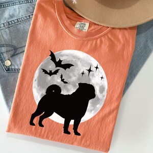 K&ouml;nnte beinhalten: Melonfarbenes T-Shirt mit einer schwarzen Silhouette eines Mopses vor einem Vollmond, Flederm&auml;usen und Sternen. Das Shirt ist auf einer Jeans und einem beigen Hut gefaltet.