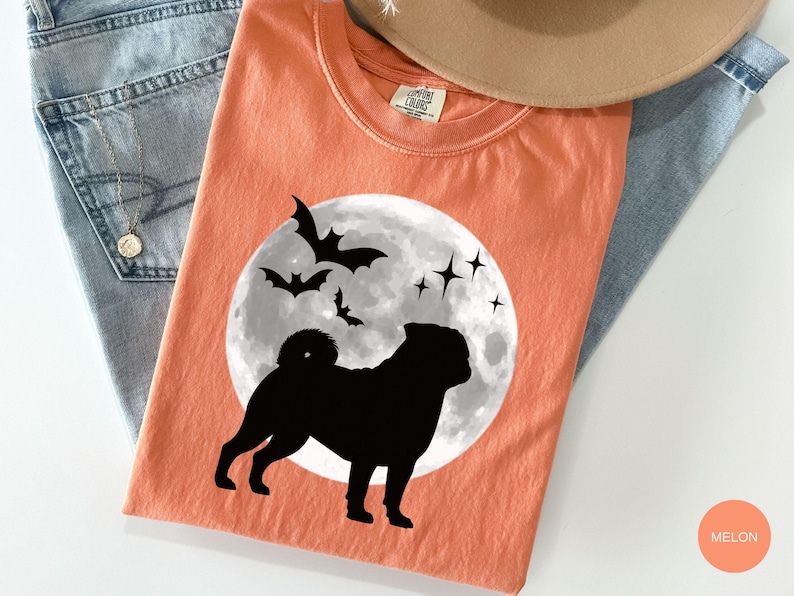 K&ouml;nnte beinhalten: Melonfarbenes T-Shirt mit einer schwarzen Silhouette eines Mopses vor einem Vollmond, Flederm&auml;usen und Sternen. Das Shirt ist auf einer Jeans und einem beigen Hut gefaltet.