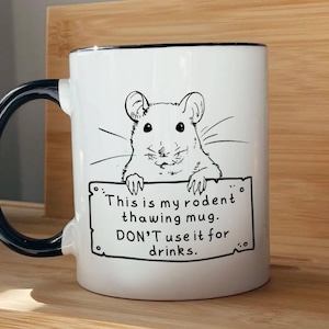 May include: White ceramic mug with a black handle and rim. The mug features a black and white illustration of a rodent holding a sign that reads, "This is my rodent thawing mug. DON'T use it for drinks."
