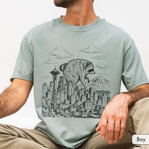 Pu&ograve; includere: Una t-shirt verde salvia con una stampa grafica nera in stile inchiostro raffigurante un procione gigante che cammina sopra lo skyline di una citt&agrave;. L'illustrazione include lo Space Needle, vari grattacieli, nuvole e una montagna.