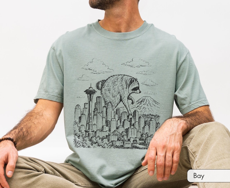 Pu&ograve; includere: Una t-shirt verde salvia con una stampa grafica nera in stile inchiostro raffigurante un procione gigante che cammina sopra lo skyline di una citt&agrave;. L'illustrazione include lo Space Needle, vari grattacieli, nuvole e una montagna.
