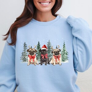 May include: Light blue sweatshirt with a Christmas-themed design featuring three pugs wearing Santa hats and reindeer antlers in a snowy forest setting.