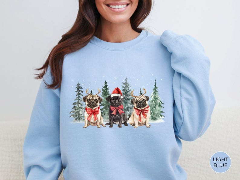 May include: Light blue sweatshirt with a Christmas-themed design featuring three pugs wearing Santa hats and reindeer antlers in a snowy forest setting.