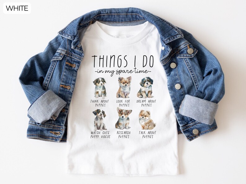 Op de afbeelding: Wit T-shirt met een blauw spijkerjasje. Het shirt heeft de tekst "THINGS I DO - in my spare time" en illustraties van puppy's. De activiteiten zijn: nadenken over puppy's, zoeken naar puppy's, dromen over puppy's, schattige puppyvideo's bekijken, puppy's onderzoeken en over puppy's praten.
