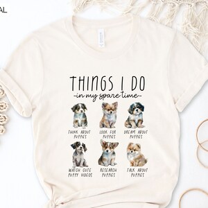 Op de afbeelding: Cr&egrave;mekleurig T-shirt met de tekst "THINGS I DO - in my spare time" en illustraties van puppy's. Het shirt heeft zes verschillende puppyrassen met bijschriften zoals "Think About Puppies" en "Watch Cute Puppy Videos".