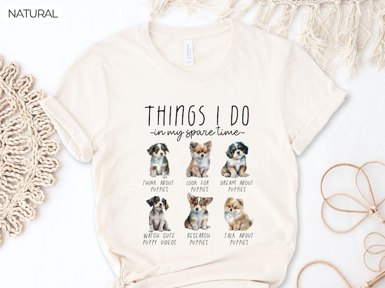 Op de afbeelding: Cr&egrave;mekleurig T-shirt met de tekst "THINGS I DO - in my spare time" en illustraties van puppy's. Het shirt heeft zes verschillende puppyrassen met bijschriften zoals "Think About Puppies" en "Watch Cute Puppy Videos".