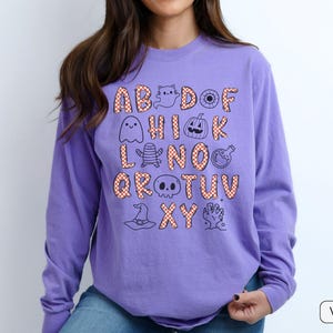 May include: A long-sleeve violet sweatshirt featuring a graphic print of the alphabet. Each letter is filled with an orange and white checkered pattern. Interspersed among the letters are black line-art illustrations of Halloween icons.