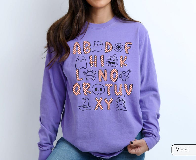 May include: A long-sleeve violet sweatshirt featuring a graphic print of the alphabet. Each letter is filled with an orange and white checkered pattern. Interspersed among the letters are black line-art illustrations of Halloween icons.