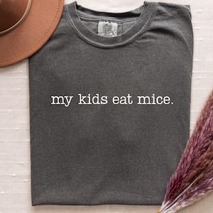 May include: Dark grey t-shirt with the text "my kids eat mice." A brown hat and a bundle of dried purple flowers are in the background. The shirt is made of a soft material.