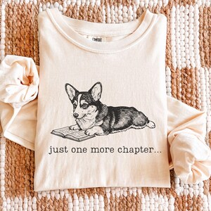 Op de afbeelding: Ivoor lange mouwen shirt met een zwart-wit illustratie van een Corgi hond die een boek leest. De tekst "just one more chapter..." is eronder gedrukt. Het shirt is op een geweven oppervlak gevouwen.