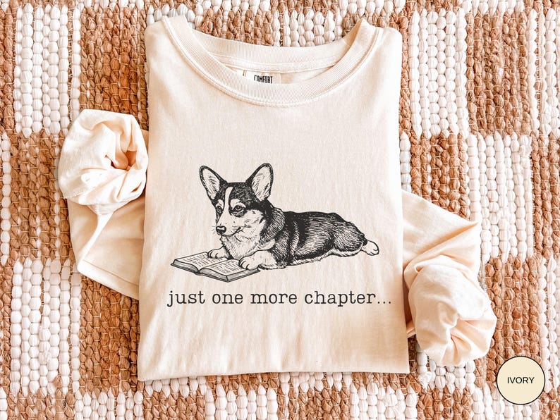 Op de afbeelding: Ivoor lange mouwen shirt met een zwart-wit illustratie van een Corgi hond die een boek leest. De tekst "just one more chapter..." is eronder gedrukt. Het shirt is op een geweven oppervlak gevouwen.