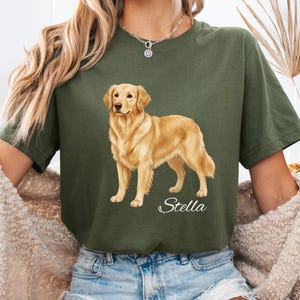 May include: Olive green t-shirt featuring a golden retriever illustration. The dog is standing, with a light brown coat and a friendly expression. The name "Stella" is written in cursive below the dog. The shirt is a relaxed fit.