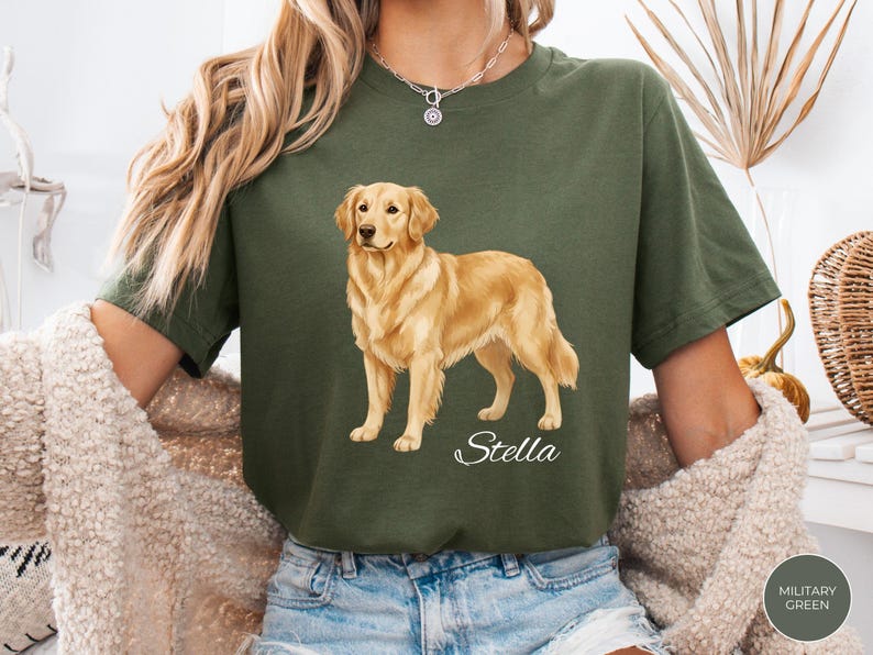 May include: Olive green t-shirt featuring a golden retriever illustration. The dog is standing, with a light brown coat and a friendly expression. The name "Stella" is written in cursive below the dog. The shirt is a relaxed fit.