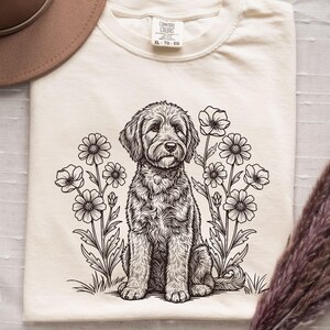 Pode incluir: Camiseta de cor marfim com uma ilustra&ccedil;&atilde;o em preto e branco de um c&atilde;o rodeado por flores. A camiseta tem gola redonda e mangas curtas. A etiqueta mostra "Comfort Colors" e o tamanho "XL-TG-EG".