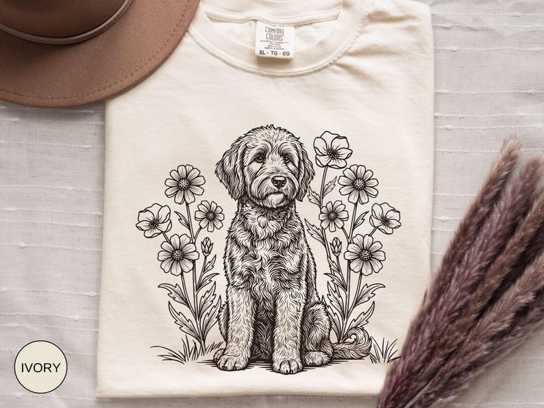 Pode incluir: Camiseta de cor marfim com uma ilustra&ccedil;&atilde;o em preto e branco de um c&atilde;o rodeado por flores. A camiseta tem gola redonda e mangas curtas. A etiqueta mostra "Comfort Colors" e o tamanho "XL-TG-EG".