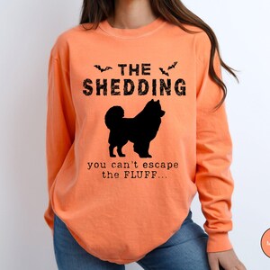 May include: Melon-colored long-sleeve shirt with the text "THE SHEDDING" above a black silhouette of a dog. Below the dog, the text reads "you can't escape the FLUFF...". Two bat silhouettes are above the word "SHEDDING".
