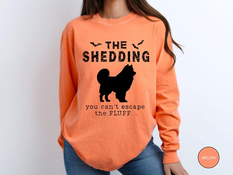 May include: Melon-colored long-sleeve shirt with the text "THE SHEDDING" above a black silhouette of a dog. Below the dog, the text reads "you can't escape the FLUFF...". Two bat silhouettes are above the word "SHEDDING".
