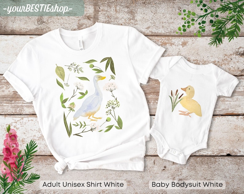 May include: A white adult unisex t-shirt and a white baby bodysuit with a watercolor illustration of a duck and a goose. The goose is surrounded by green leaves and white flowers. The duck is surrounded by green reeds. The text "yourBESTIEShop" is at the top of the image.